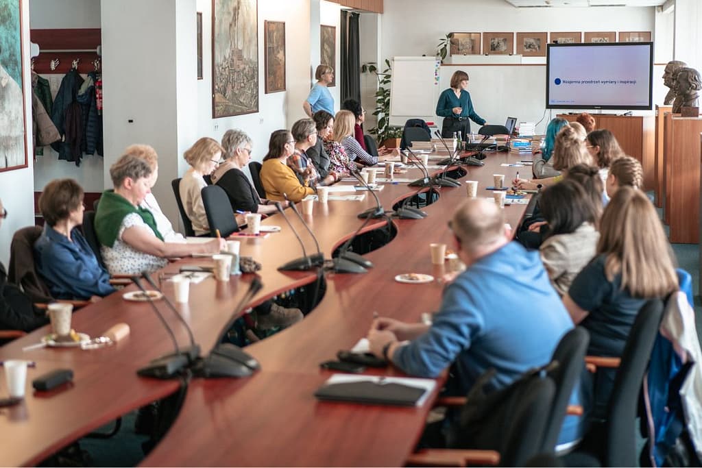 Grupa osób siedzi przy długim, zakrzywionym stole konferencyjnym podczas prezentacji. W tle kobieta stoi przy ekranie prowadząc spotkanie dla uczestników.