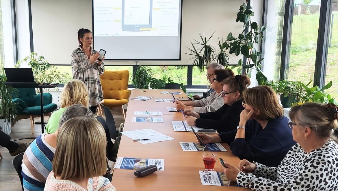 Seniorzy siedzą w sali warsztatowej, prowadząca pokazuje czytnik ebooków i omawia jego funkcje.