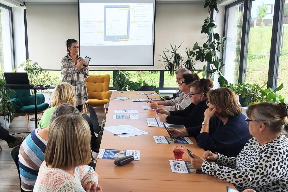 Warsztaty dla seniorów z obsługi czytników ebooków w bibliotece Seniorzy siedzą w sali warsztatowej, prowadząca pokazuje czytnik ebooków i omawia jego funkcje.