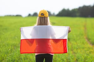 Dziewczynka trzymająca za plecami czerwoną flagę patrzy na zieloną łakę.