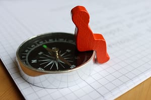 Zbliżenie na metalowy kompas turystyczny leżący na kartce papieru w kratkę. Na krawędzi kompasu siedzi mała, drewniana, czerwona figurka przypominająca człowieka.